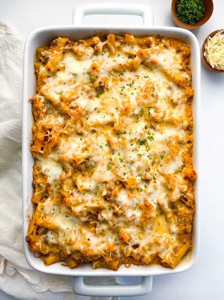A baking dish filled with baked ziti, near a serving napkin, extra parsley, and shredded parmesan cheese. Healthy Food, Real Food, Real Food Recipes, Healthy Food Recipes, Wholesome food, Wholesome food recipes, Whole Food, Whole Food Recipes, Realgood food, Realfooddaily Eat real, Good Real Food, Goodrealfood, baked ziti, pasta, ground beef, casserole, baked ziti pasta, baked noodles, marinara sauce, easy baked dinners, ziti noodle