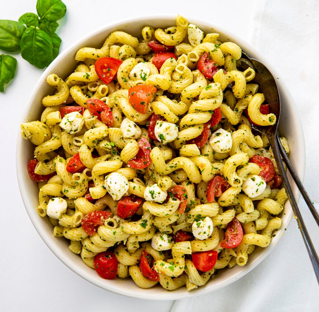 A bowl of caprese pesto pasta salad with utensils and a cluster of basil. Healthy Food, Real Food, Real Food Recipes, Healthy Food Recipes, Wholesome food, Wholesome food recipes, Whole Food, Whole Food Recipes, Realgood food, Realfooddaily Eat real, Good Real Food, Goodrealfood, pesto, pesto pasta, pasta, caprese pasta, caprese pesto pasta, tomato pasta, pine nuts, cold pasta salad, easy pasta, side dish,