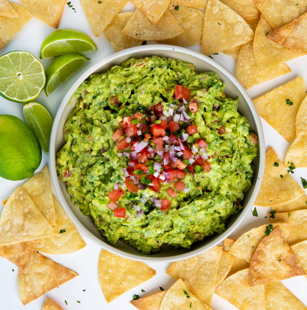 A serving bowl of guacamole, topped with tomatoes and onions, surrounded by chips and limes. Healthy Food, Real Food, Real Food Recipes, Healthy Food Recipes, Wholesome food, Wholesome food recipes, Whole Food, Whole Food Recipes, Realgood food, Realfooddaily Eat real, Good Real Food, Goodrealfood, guacamole, healthy dip, fresh dip, easy dip, fresh veggie dip, vegetable dip, avocado dip, appetizer, side dish