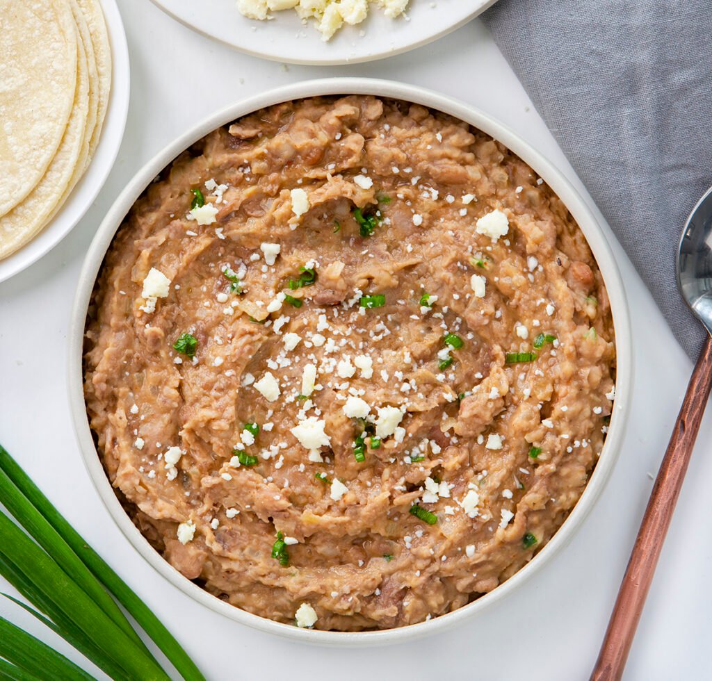 A bowl of refried beans, near a plate of tortillas, queso fresco cheese, green onions, a towel and a serving spoon. Healthy Food, Real Food, Real Food Recipes, Healthy Food Recipes, Wholesome food, Wholesome food recipes, Whole Food, Whole Food Recipes, Realgood food, Realfooddaily Eat real, Good Real Food, Goodrealfood, refried beans, homemade refried beans, beans, bean recipes, pinto beans, fresh refried beans, how to make beans from scratch, from scratch cooking,