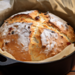 A loaf of rustic sun dried tomato bread in a black dutch oven with brown parchment paper.