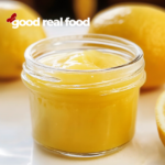 A small jar of lemon curd, with fresh lemons in the background.