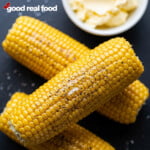 Corn on the cob, seasoned with salt and pepper, a bowl of butter rests in the background.