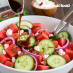 A white bowl filled with Watermelon Salad. Balsamic Dressing is being poured over the salad.