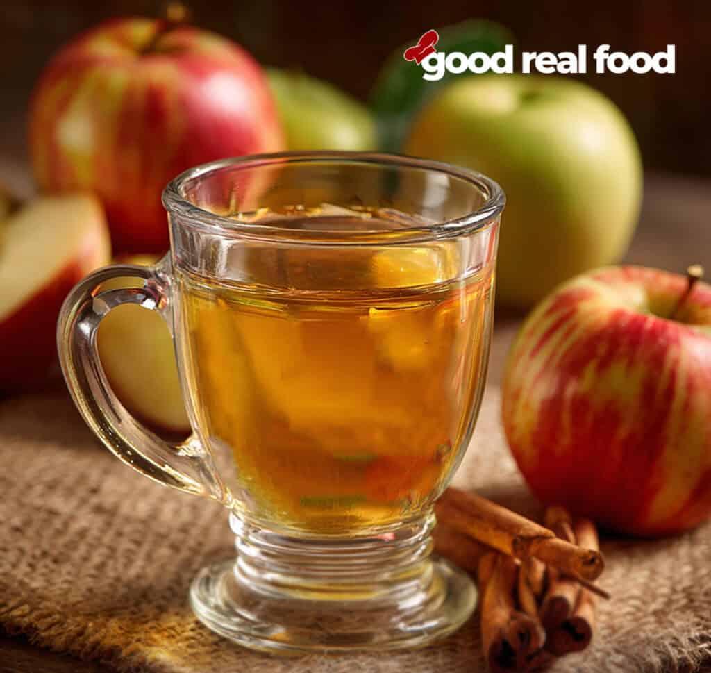 A clear glass of homemade apple cider surrounded by apples and cinnamon sticks.