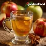 A clear glass of homemade apple cider surrounded by apples and cinnamon sticks.