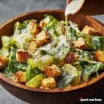 A wooden bowl filled with a classic Caesar Salad. Caesar dressing is being poured over the salad.