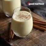 A glass of eggnog with cinnamon sticks on a wooden surface.