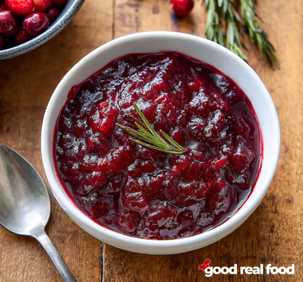 A white bowl of easy cranberry sauce rests on a wooden surface with a sprig of rosemary.