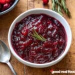 A white bowl of easy cranberry sauce rests on a wooden surface with a sprig of rosemary.