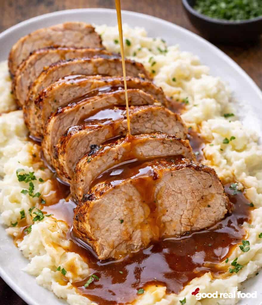 Maple Mustard Pork Tenderloin served over mashed potatoes on a white platter. Sauce is being drizzled over the meat.