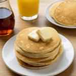 Stack of Fluffy Buttermilk Pancakes on a white plate on a wood table with syrup and orange juice and another plate of pancakes in the background.