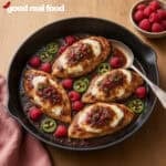 a cast iron pan of raspberry jalapeno cream cheese chicken with a spoon on a wood counter next to a cloth napkin and a little bowl of raspberries