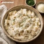 close up Creamy Slow Cooker Chicken Alfredo in a ceramic on a wood counter next to a cloth napkin and two small bowls with parmesan and parsley
