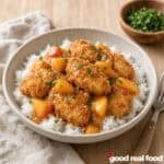 A bowl of crispy coconut peach chicken over rice on a wood counter next a fork and a cloth napkin