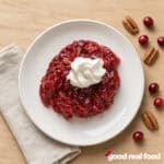 Holiday cranberry salad with whipped cream on a white plate on a wood counter next to a cloth napkin and cranberries and pecans