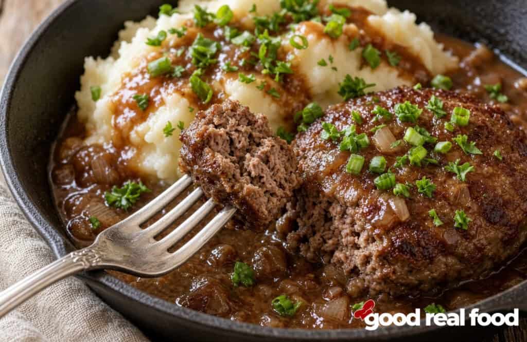 salisbury steak on a fork over pan of salisbury steak with onion gravy and mashed potatoes a close up of a bite of salisbury steak on a fork over a pan of salisbury steak with onion gravy and mashed potatoes next to a cloth napkin
