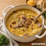 a white pot of spicy cajun potato soup next to a cloth napkin, a small bowl of cheese, and some bread, with garnish