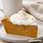 a slice of crustless pumpkin pie with whipped cream on a stone plate on a wood counter with napkin and coffee mug in the background