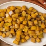 Maple Cinnamon Roasted Squash on a white plate on a wood counter next to a bowl of garnish and a cloth napkin