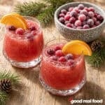 two glasses of Orange Cranberry Slush garnished with sugared cranberries and orange slices on a rough wood counter next to a bowl of sugared cranberries some pine tree greenery