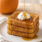 a stack of pumpkin spice French toast with syrup being drizzled on top on a white plate on a cloth napkin on a wood counter next to a pumpkin